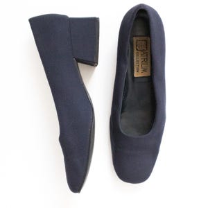 May include: A pair of navy blue low-heeled shoes. The shoes have a rounded toe and a block heel. The interior of one shoe has a label that reads "ATRIUM COLLECTION". The shoes are made of a textured fabric.