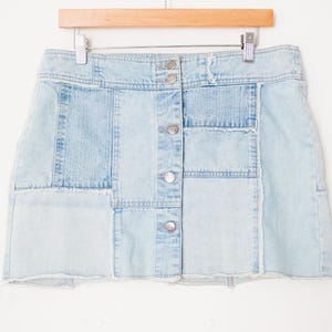 May include: Light blue denim skirt with a patchwork design. The skirt features a button-up front and a raw hem. The skirt is hanging on a wooden hanger against a white background. The skirt is made of various shades of light blue denim.