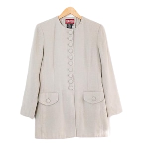 May include: A light beige long coat with a round collar and long sleeves. The coat has a row of buttons down the front, two pockets, and a small label at the neckline. The coat is hanging on a wooden hanger against a white background.