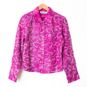 Puede incluir: Chaqueta rosa brillante con botones y un patrón blanco en espiral. La chaqueta tiene mangas largas y cuello. La etiqueta dice "AMAEE". Cuelga de una percha de madera sobre un fondo blanco.
