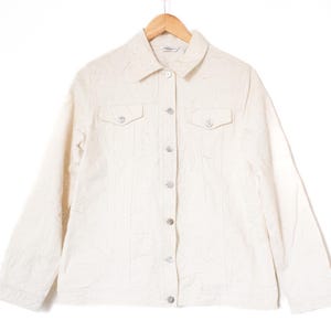 May include: Cream-coloured jacket with a collared neckline, two chest pockets, and silver buttons. The jacket hangs on a wooden hanger. The fabric has a subtle textured pattern. A classic, casual outerwear piece.