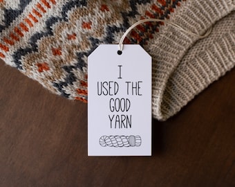 May include: A white rectangular paper tag with a string, featuring the black printed text I USED THE GOOD YARN and a small illustration of a yarn skein, resting on a beige knit garment with a geometric pattern in orange, gray, and white.