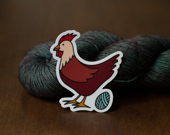 May include: A white sticker with a cartoon illustration of a red rooster standing on a ball of yarn. The yarn is teal and black.