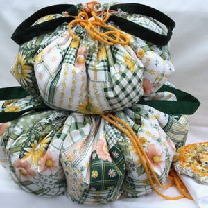 Könnte beinhalten: Zwei gestapelte Stoffbeutel mit Kordelzug und einem Patchwork-Muster aus gelben Sonnenblumen, grünem Karomuster und Blumenmotiven. Jeder Beutel hat dunkelgrüne Stoffgriffe und einen leuchtend orangefarbenen Kordelzug. Daneben liegt eine kleine, strukturierte, runde Matte.