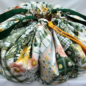 Könnte beinhalten: Ein runder Stoffbeutel mit Kordelzug, der ein Patchwork-Design aus weißen, grünen und gelben Blumen- und Karomustern aufweist. Die Tasche verfügt über dunkelgrüne Stoffgriffe und einen leuchtend gelben Kordelzug, der die obere Öffnung zuzieht.