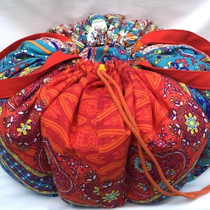 Könnte beinhalten: Ein runder Stoffbeutel mit Kordelzug, der mit einem lebendigen, mehrfarbigen Paisley- und Blumenmuster in Rot-, Orange-, Blau- und Gelbtönen versehen ist. Die Tasche verfügt über zwei einfarbige rote Stoffgriffe und einen orangefarbenen Kordelzug.