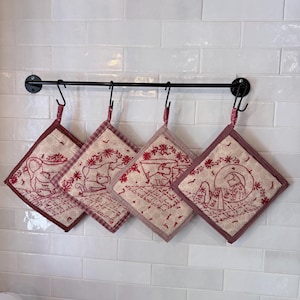 May include: Four square quilted potholders with red embroidered animal illustrations on a cream background, each featuring a red checkered border and a fabric loop, hanging from a black metal rod with S-hooks in a row against a white tiled wall.