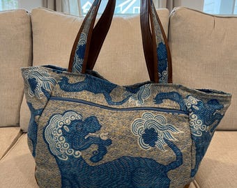 May include: A blue and beige tote bag with a brown leather handle. The bag features a repeating pattern of blue tigers with white clouds.