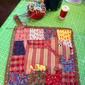 May include: A handmade quilted hot pad featuring a patchwork design with a central appliqué of a Scottish Terrier dog. The pad is made of various fabrics in red, blue, and brown tones, with a brown border. A charming and functional kitchen accessory.
