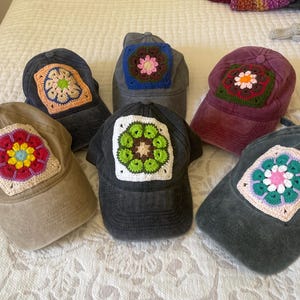 Peut inclure: Plusieurs casquettes de baseball de couleurs variées, chacune ornée d'un motif carré crocheté unique et coloré. Les casquettes sont dans des tons de gris, marron et bordeaux, avec des motifs floraux crochetés.