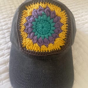 May include: A charcoal grey baseball cap featuring a square crochet patch on the front panel. The patch displays a circular floral pattern with a teal centre, surrounded by layers of purple and yellow yarn, all bordered by a dark brown crochet frame.