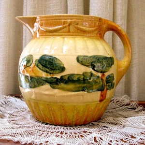 May include: A yellow and green glazed ceramic pitcher with a handle. The pitcher has a floral design with green leaves and yellow flowers. The pitcher has a ribbed design and a brown rim.