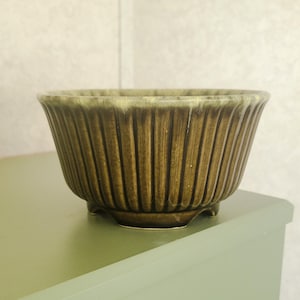 May include: A brown ceramic bowl with vertical ribbed detailing. The bowl has a rounded shape and sits on three small feet. The rim has a lighter, yellowish-green glaze. The bowl is likely vintage and suitable for decorative or functional use.