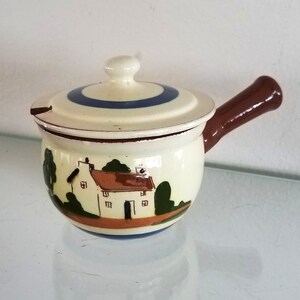 May include: A small white ceramic creamer with a brown handle and a blue and white striped lid. The creamer is decorated with a painted scene of a white house with a brown roof and green trees.
