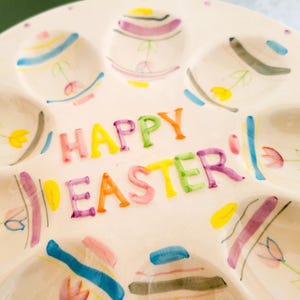 May include: A white ceramic Easter egg serving platter with six egg-shaped indentations. The platter is decorated with colorful painted Easter eggs and the words "HAPPY EASTER" in bright, playful lettering. The eggs are painted with pastel colors and floral accents.