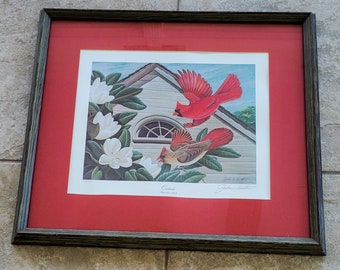 May include: Framed print featuring two cardinals, one red and one brown, flying near a white house with a curved window and white magnolia flowers. The print is set against a red mat and framed in a dark wood.