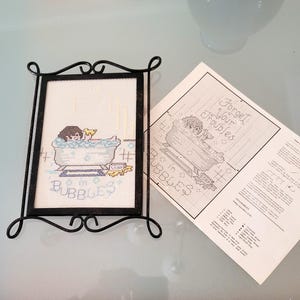 May include: A framed cross-stitch piece depicting a person in a bathtub with the word "Bubbles" below. The frame is black metal with decorative swirls. A pattern sheet with the text "Forget Your Troubles" is next to the frame.