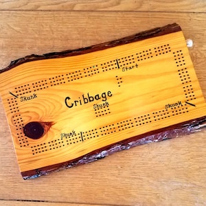 May include: A wooden cribbage board with a natural wood finish. The board has a unique shape and is marked with black dots and the words "Start", "Skunk", and "Cribbaage".
