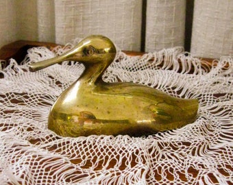 May include: A brass duck figurine with a long beak and a smooth, reflective surface. The duck is resting on a white, open-weave doily. The figurine is a decorative item.