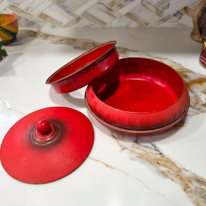 May include: A round, bright red metal container with a matching lid featuring a small central knob. The container consists of a deep base and a separate, slightly smaller circular rim piece, both finished in a vibrant, solid red color.