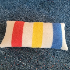 May include: A rectangular pillow with a white background and three vertical stripes in red, yellow, and blue. The pillow has a soft, textured appearance, suggesting a wool or similar fabric. The design is simple and colorful.