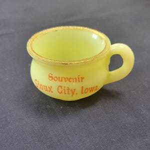 May include: A yellow glass souvenir cup with a gold rim and the words "Souvenir Sioux City, Iowa" printed in orange.