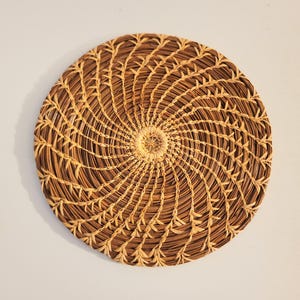 May include: A round, woven basket with a spiral design. The basket is made of natural brown and tan materials, with a central circular pattern. The spiral pattern radiates outwards from the center, creating a textured surface. The basket is likely used for decorative or functional purposes.