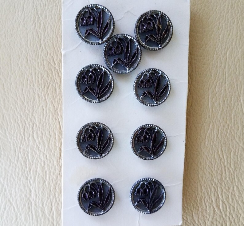 May include: A pack of ten black and silver buttons with a floral design. The buttons are round and have a raised design of a flower with a stem and leaves.
