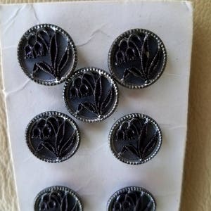 May include: A set of eight black and silver buttons with a floral design. The buttons are round and have a raised design of a flower with a stem and leaves.