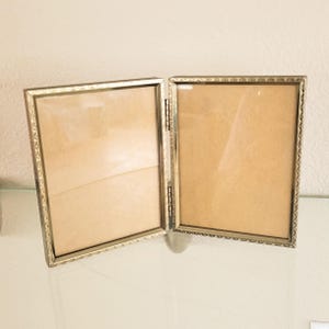 May include: A double-hinged metal picture frame with a gold-toned finish and a textured, repeating geometric pattern along the outer edges. The frame features two rectangular openings for photographs, connected by a central vertical metal hinge.