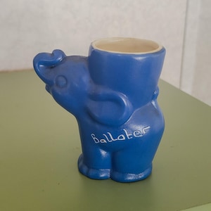 May include: A blue ceramic elephant-shaped container with a cream-colored interior. The word "Ballater" is written in cursive on the side. The container is standing on a green surface.