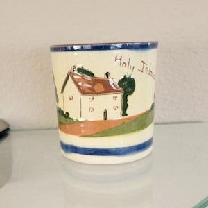 May include: A cylindrical ceramic mug with a cream-colored base featuring a hand-painted scene of a house with a brown roof, green trees, and a path. The text Holy Island is written in brown script. The mug has dark blue bands at the top and bottom.