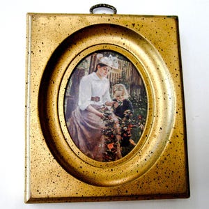 May include: A gold-toned oval picture frame with a print of a woman and a child in a garden setting. The woman is wearing a white blouse and a straw hat. The child is wearing a dark dress. The frame has a textured surface.