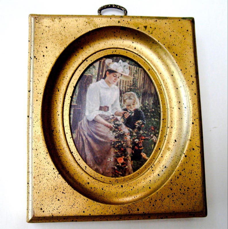 May include: A gold-toned oval picture frame with a print of a woman and a child in a garden setting. The woman is wearing a white blouse and a straw hat. The child is wearing a dark dress. The frame has a textured surface.