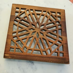 May include: Square wooden coaster with a geometric, openwork design. The coaster is made of a warm-toned wood and features a symmetrical pattern of intersecting lines and shapes. The coaster is approximately 5 inches square.