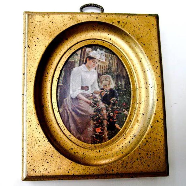 May include: A gold-toned picture frame with a raised oval border. The frame contains a vintage print of a woman and a child in a garden setting.