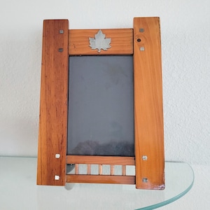 May include: A rectangular wooden picture frame with a dark brown finish, featuring a vertical orientation. The frame includes a silver-colored metal maple leaf emblem at the top center and small square metal accents on the side panels.