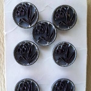 May include: Eight silver-colored buttons with a black lily of the valley design. The buttons are round and have a raised edge.