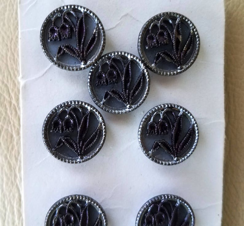 May include: Eight silver-colored buttons with a black lily of the valley design. The buttons are round and have a raised edge.