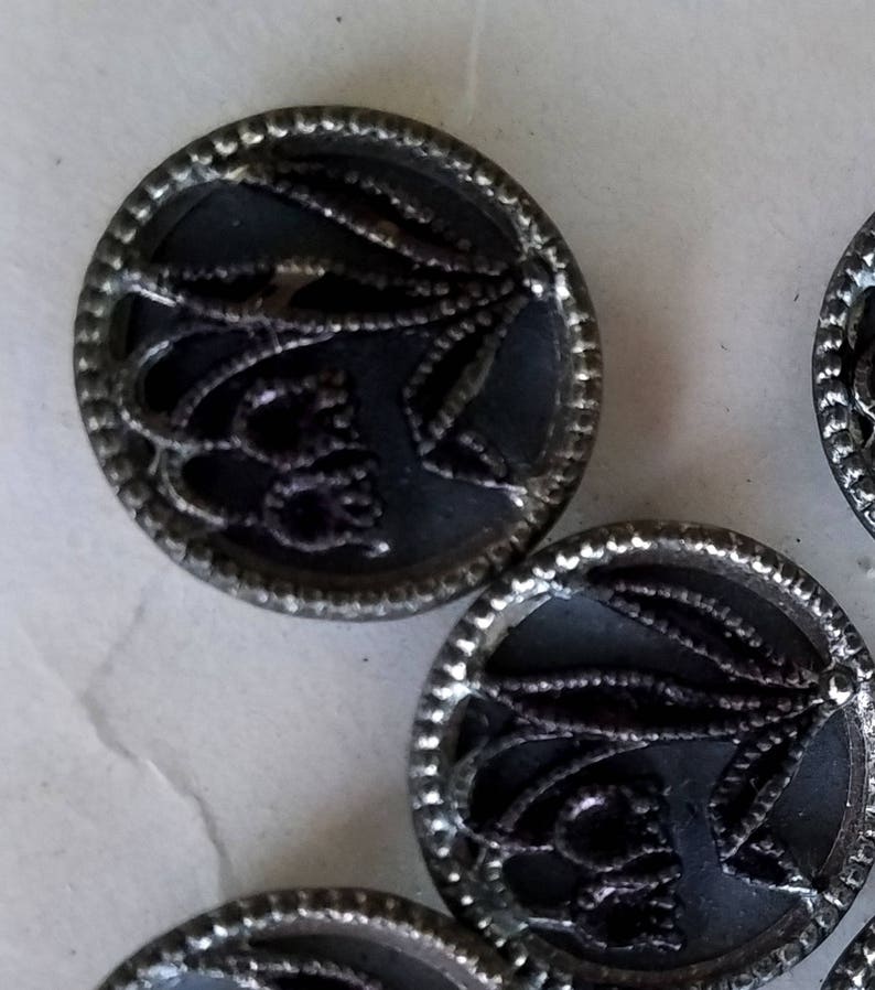 May include: A set of six silver-toned buttons with a black, intricate design. The buttons have a raised, circular rim and a flat center.