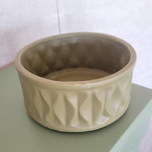 May include: A round, olive-green ceramic bowl with a textured diamond pattern around the exterior. The bowl has a smooth interior and sits on a green surface. The bowl is a decorative item.