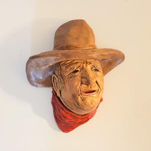 May include: A sculpted wall hanging depicting a weathered face of a man wearing a brown cowboy hat and a red scarf. The sculpture has detailed facial features and is mounted on a white wall.