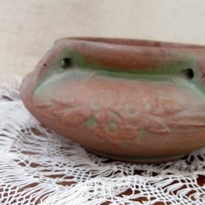May include: A small, brown ceramic planter with a green accent band and floral design. The planter has two small holes and sits on a white lace surface. The overall design is rustic and vintage.