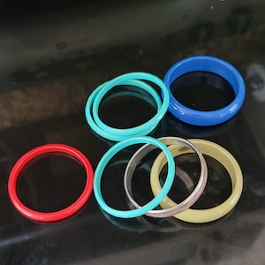 May include: A collection of colorful bangles in various shades of red, blue, green, and yellow. The bangles are made of different materials, including plastic and metal, and are arranged on a reflective black surface.