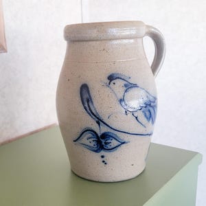 May include: A stoneware pitcher with a light beige glaze and a blue bird and leaf design. The pitcher has a rounded body, a handle, and a slightly flared rim. The bird is depicted with simple lines and shading, and the leaves are stylized.