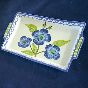 May include: Rectangular ceramic serving tray with a white base, adorned with blue floral designs and green leaves. The tray has a blue scalloped border and green accents on the handles. A decorative piece for serving.