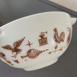 May include: White mixing bowl with brown illustrations of a rooster, eagle, coffee grinder, and other kitchen-themed designs. The bowl has a handle and a rounded shape, perfect for mixing ingredients.