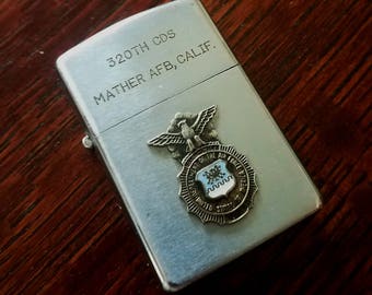 May include: Silver lighter with the inscription "320TH CDS MATHER AFB, CALIF." and a silver emblem of the United States Air Force.