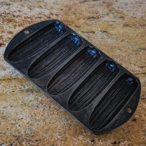 May include: A black cast iron cornbread pan with five individual molds. The molds are shaped like corn cobs and have a textured surface.