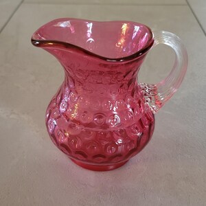 May include: A small, pink glass pitcher with a clear glass handle. The pitcher has a textured, bubble-like design.
